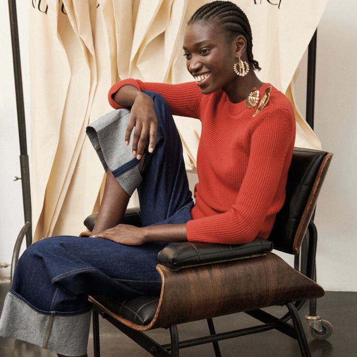 New Patou collection, presenting in a photoshoot with a model seating on a chair wearing denim pants and red pullover