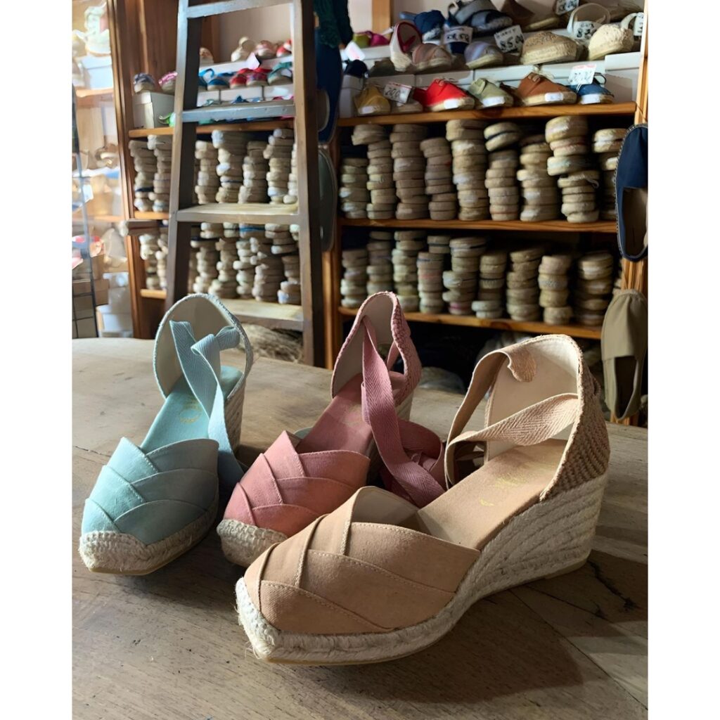 close up of three model of wedge Espadrilles on a counter in front of a shelf full of other espadrilles models