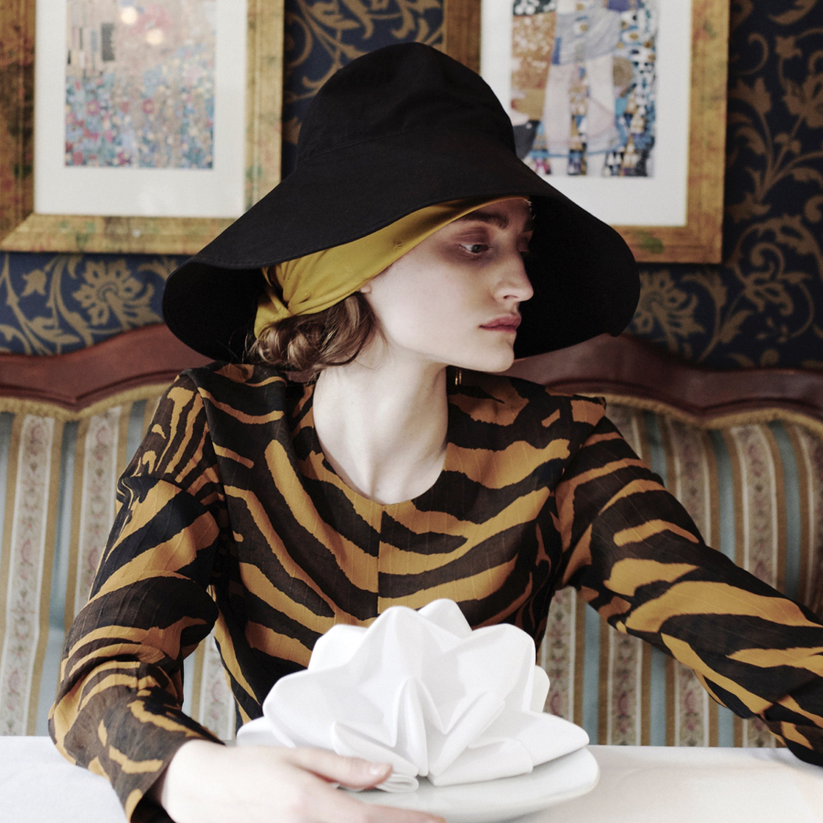 Model sitting at a train bar, wearing a zebra-print dress, hat and silk scarf.