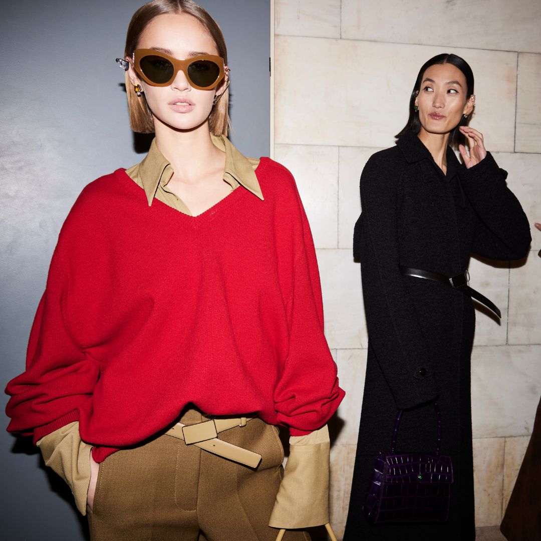 two models in the backstage of TOry Burch AW 2023 runway show. One is wearing a red pullover over a camel satin blouse and camel trousers and the other one is wearing a black overcoat.