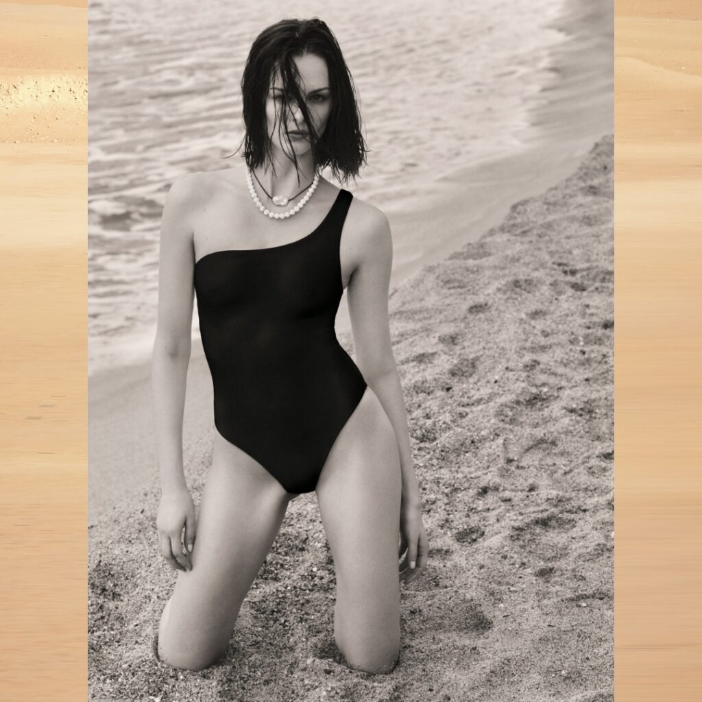 model wearing a black swimsuit and pearl necklace is kneeling on the beach with a wet hair