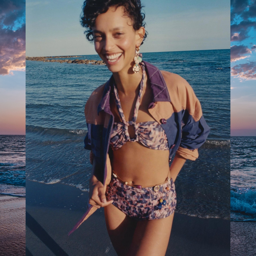 woman wearing a SESSUN bikini and a shirt stands on the ocean's shore smiling to the camera.