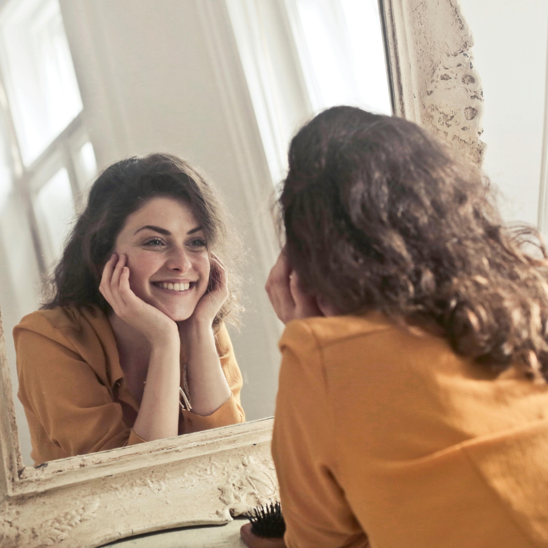 happy woman looking at her skin in a mirror standing on a fireplace