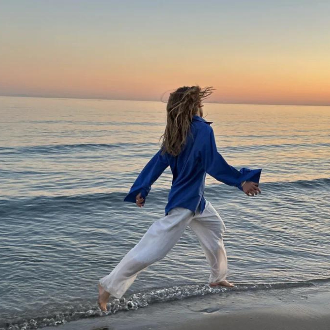 model walking along the water's edge at the seaside during sunset. She is wearing white pants and a cobalt blue shirt.