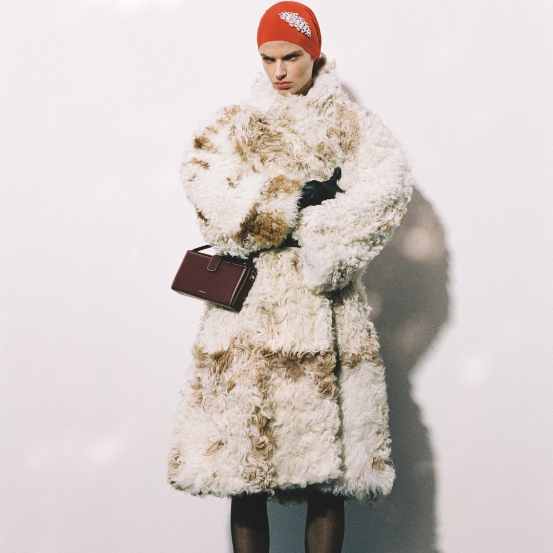 Magda Butrym model wearing a faux fur coat and a red beanie with a crystal brooch looks at the camera