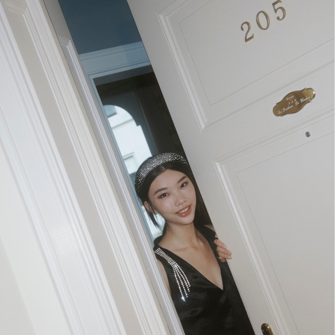 A model wearing a black dress and a sequined hairband is opening the door of a hotel room, looking inviting to the camera