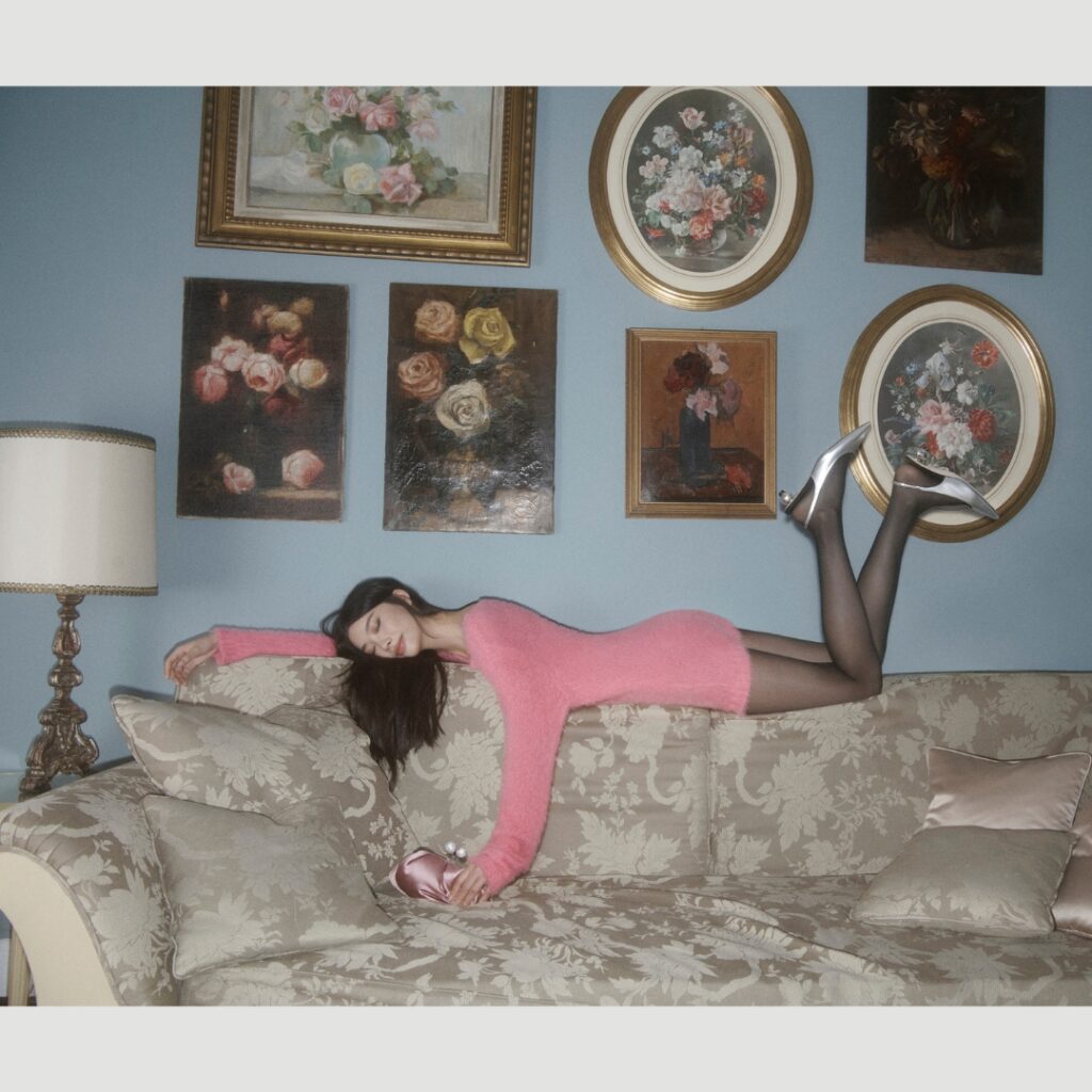 a model laying on the top of a sofa in a hotel room, wearing a knit pink dress and a silver back sling pump