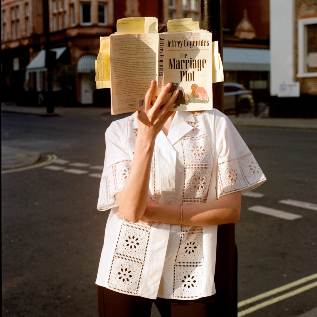 Model wearing a lines embroidered white shirt stands on the street with an open book in front of her face
