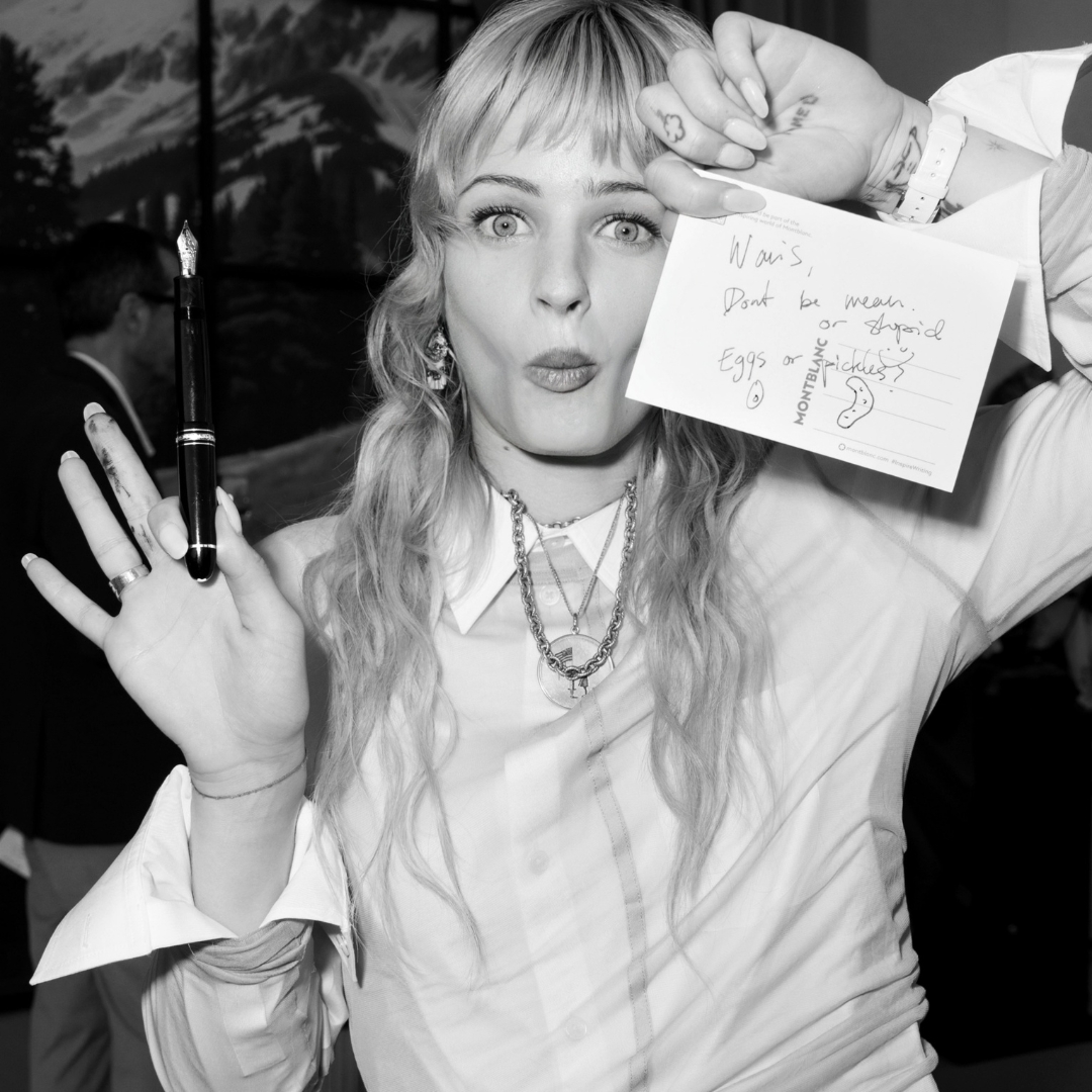Esther MacGregor in a black and photo from German Larkin-She shows to the camera a postcard while holding a Montblanc pen