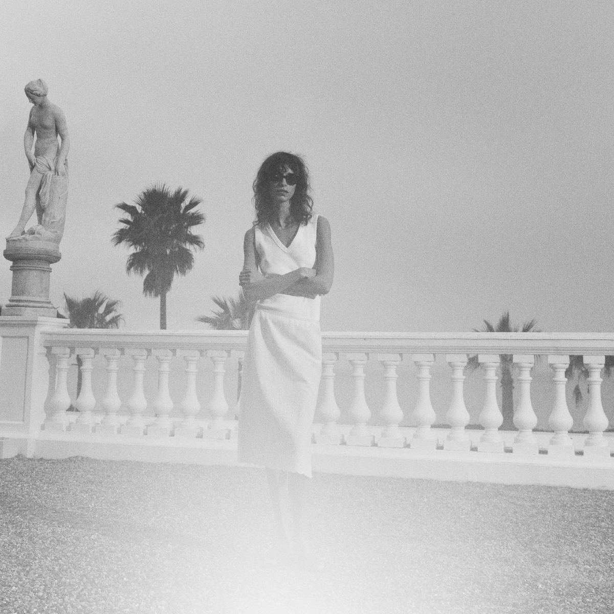 woman wearing a white outfit, standing on a terrace with palm trees and the ocean in the background