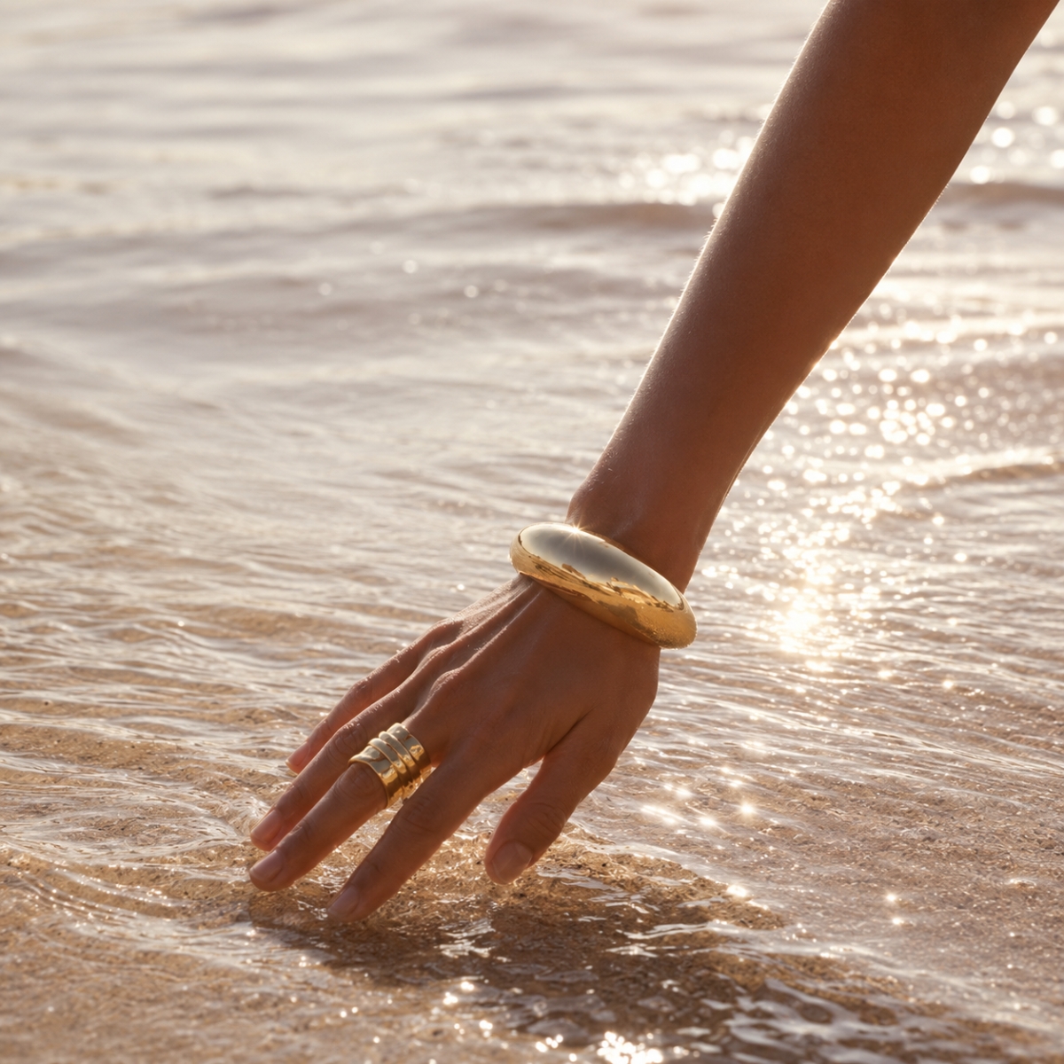 Female arm wearing a gold cuff and a gold ring caresse the sea water on the shore